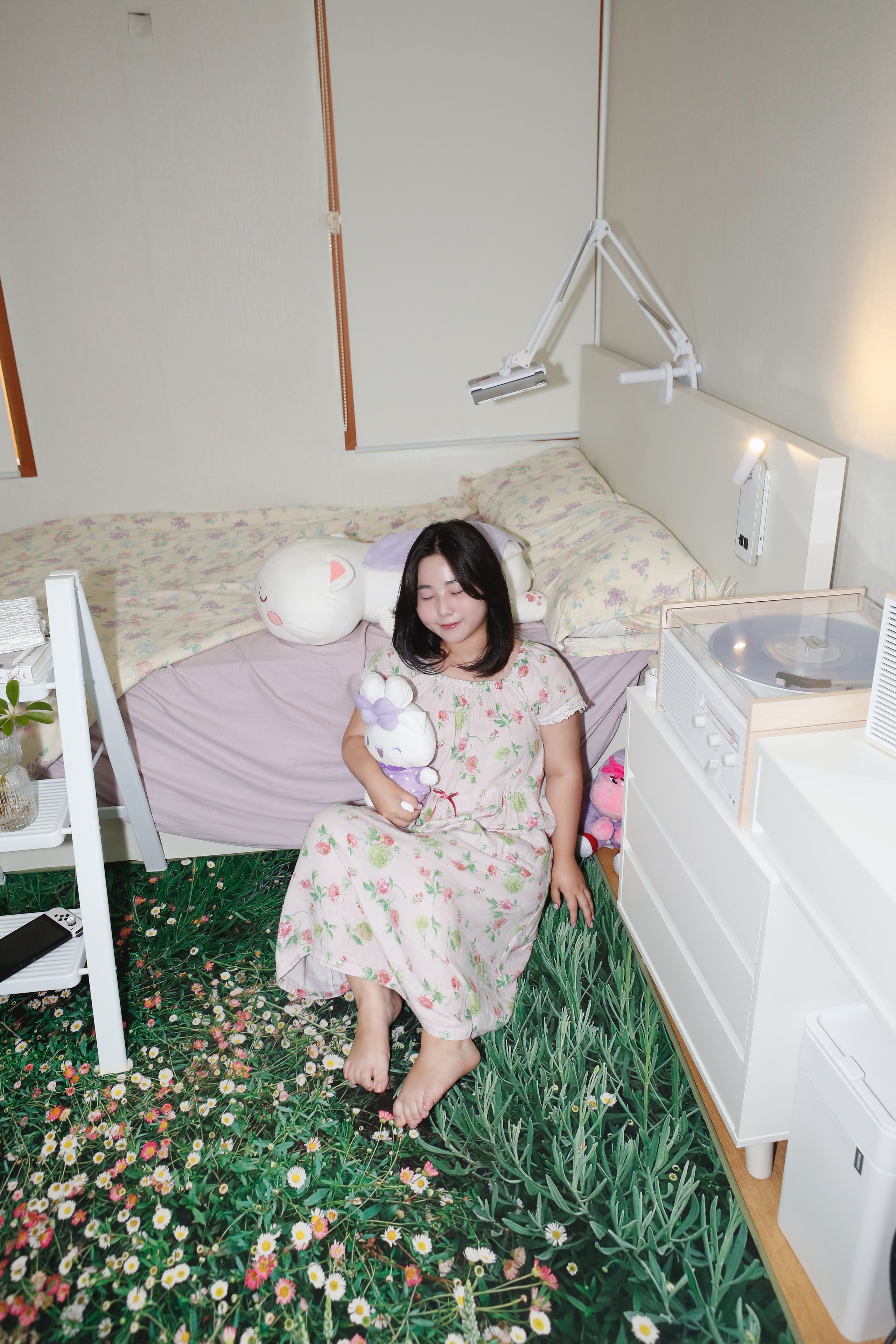 Woman in a pink floral dress sits on a flower-patterned bed holding a white plush toy in a bedroom.